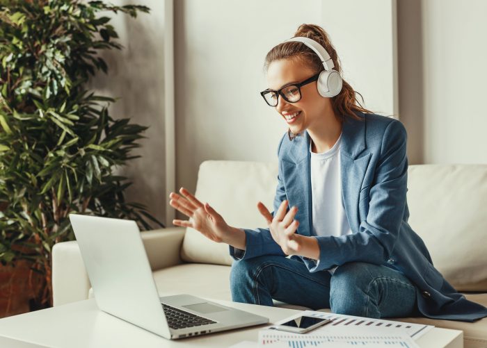 Cheerful young female in casual outfit and glasses having video conversation with colleagues and sharing business ideas while sitting on couch in modern workplace